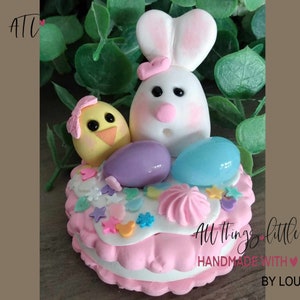 May include: A pink and white cake decorated with pastel colored fondant, sprinkles, and a bunny and chick figurine. The cake is decorated with a pink and white swirl and the text "All Things Little Handmade With Love By Lou".