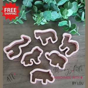 May include: Pink plastic animal-shaped cookie cutters. The set includes a bear, lion, elephant, hippo, and other animal shapes. The text "All Things Little Handmade with Love by Lou" is visible.