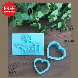 May include: A light blue cookie stamp with the words "I love you" and two heart-shaped cookie cutters. The stamp is rectangular and the text is in a cursive font. The cookie cutters are in the shape of a heart.