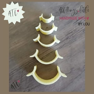 May include: Five pale yellow, crescent-shaped objects arranged in a vertical line from smallest to largest. Each piece features a curved base with flared, wing-like ends. The text "ATL" and "All things little HANDMADE WITH BY LOU" is visible.
