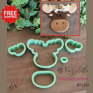May include: A brown moose-shaped cookie with a smiling face and decorative antlers, displayed with green plastic cookie cutters in the shape of a moose, heart, and oval. The text "All things little" is visible.