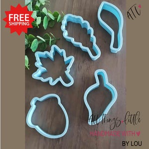 May include: Set of five blue plastic cookie cutters shaped like an acorn, a leaf, a sprig of leaves, a bell pepper, and a maple leaf. The cutters are on a wooden surface. All Things Little Handmade with Love by Lou.
