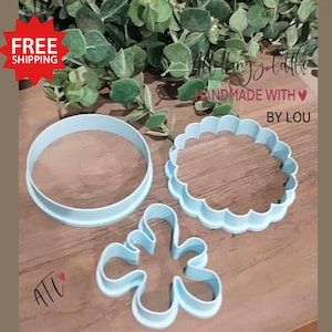 May include: Three blue plastic cookie cutters: a circle, a scalloped circle, and a flower shape. The cutters are on a wooden surface. "All Things Little Handmade with Love By Lou" is written on the surface.