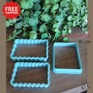 May include: Three light blue cookie cutters. Two have a scalloped edge, and one is a simple rectangle. The cookie cutters are on a wooden surface with green foliage in the background. The text "All Things Little Handmade With Love By Lou" is visible.