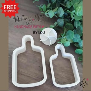 May include: Two white plastic cookie cutters shaped like bottles. The cutters are on a wooden surface with green foliage in the background. The text "All Things Little Handmade with Love By Lou" is visible in the image.