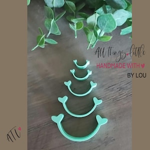 May include: Five light green, curved, crescent-shaped objects with small heart-shaped ends arranged in a vertical stack from smallest to largest. The text "All things little HANDMADE WITH BY LOU" is visible on the right side of the image.