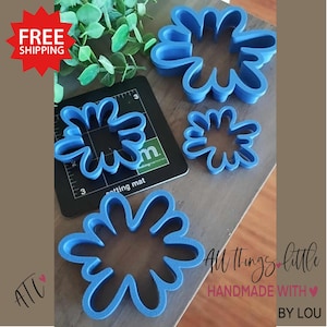 May include: Set of four blue plastic cookie cutters shaped like abstract flowers. The cutters are arranged on a wooden surface. The text "All things little" and "Handmade with" is visible in the image.