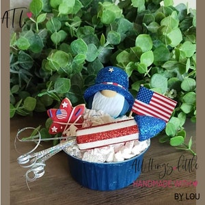 May include: A patriotic gnome wearing a blue and white hat with a star on top, sitting in a blue cup with white marshmallows and a red, white, and blue firework. The gnome is holding a small American flag. The text "All Things Little Handmade With BY LOU" is visible at the bottom of the image.