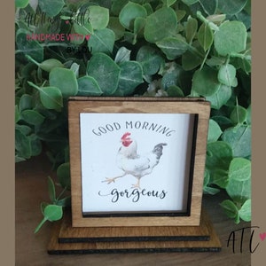 May include: Wooden sign with a white rooster illustration and the text "Good Morning Gorgeous". The sign is on a tiered wooden stand.