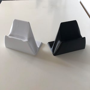 Pode incluir: Dois suportes para telefone impressos em 3D, um branco e outro preto. Os suportes são inclinados para segurar um telefone na posição vertical.