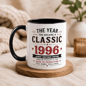 Puede incluir: Una taza de cerámica blanca con asa y borde interior negros. El frente presenta texto en negro y rojo: THE YEAR YOU BECAME A CLASSIC 1996, JAMES' BIRTHDAY EDITION, BORN ON FEBRUARY 21, 1996, AGED TO PERFECTION, STILL ICONIC, STILL LOVED.