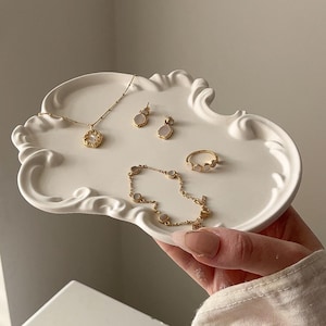 May include: A white, ornate tray holds a collection of gold-toned jewelry. The jewelry includes a necklace, earrings, a bracelet, and a ring, all featuring delicate designs and small, round, light-colored stones. The tray is held by a person.