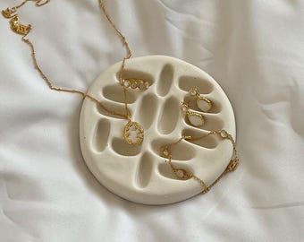 May include: A gold necklace with a teardrop pendant, a gold bracelet, and two small gold hoop earrings arranged on a circular white ceramic tray featuring a series of recessed oval indentations across its surface.