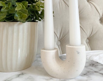 Könnte beinhalten: Ein moderner Kerzenhalter mit zwei weißen Spitzkerzen. Der Halter hat eine gesprenkelte beige Farbe mit einem geschwungenen Design. Eine grüne Topfpflanze befindet sich links von den Kerzen, und der Hintergrund ist eine getuftete, cremefarbene Oberfläche.