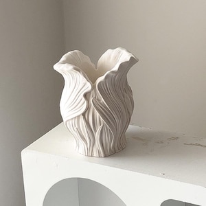 May include: A white ceramic vase with a floral design. The vase has a textured surface with vertical lines and a flared top. It is on a white shelf against a white wall.