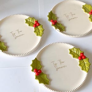 May include: Three round white ceramic plates with a beaded edge, each featuring the text 'Tis The Season' in black script and decorated with two clusters of green holly leaves and bright red berries on opposite sides of the plate rim.