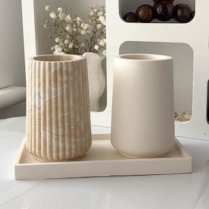 Peut inclure: Deux vases cylindriques sont posés sur un plateau rectangulaire. Le vase de gauche présente une texture nervurée verticale avec un motif marbré beige et blanc. Le vase de droite a une finition blanc cassé mate et lisse sur le plateau.