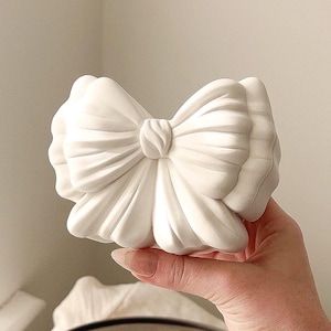 May include: A white, bow-shaped ornament. The bow is sculpted with detailed folds and a central knot. The object is held in a hand, with a neutral background.