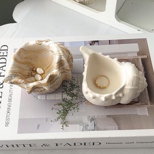 Könnte beinhalten: Zwei muschelförmige Schmuckschalen aus Keramik auf einem Buch mit dem Titel White & Faded. Eine Schale hat ein marmoriertes beige-weißes Muster, die andere ist einfarbig weiß. Ein goldener Ring liegt in der weißen Muschel, Ohrringe in der marmorierten.