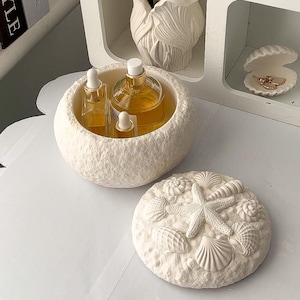 Könnte beinhalten: Eine weiße, strukturierte, runde dekorative Schale enthält drei Glasflaschen mit bernsteinfarbener Flüssigkeit. Ein passender Deckel mit Muschel- und Seestern-Designs befindet sich in der Nähe. Ein weißes Regal zeigt eine Muschel und eine Vase mit weißen Blumen.