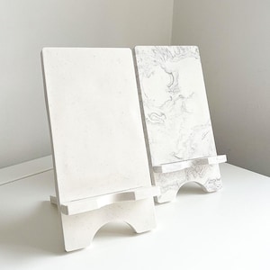 May include: Two white and gray concrete phone stands. One stand is solid white, the other has a marbled gray pattern. Both have a rectangular shape with a small ledge at the bottom to hold a phone or tablet.