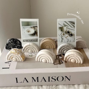 May include: A selection of rainbow-shaped ornaments in shades of white, beige, and marble, some with gold leaf detailing. The objects are displayed on a white box with the text "LA MAISON" and two framed photographs. The text "Personalise me" is also visible.