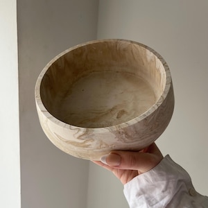 May include: A round, beige bowl with a marbled pattern. The bowl is held up, showcasing its interior and exterior design. The color palette includes shades of beige and brown, creating a natural, earthy aesthetic. The bowl appears to be made of a solid material.