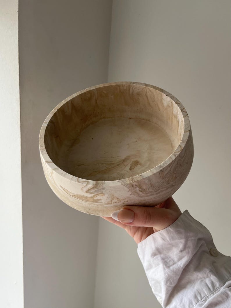 May include: A round, beige bowl with a marbled pattern. The bowl is held up, showcasing its interior and exterior design. The color palette includes shades of beige and brown, creating a natural, earthy aesthetic. The bowl appears to be made of a solid material.