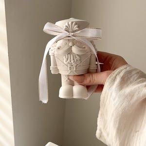 May include: A matte white nutcracker figurine crafted from a solid material, featuring detailed moulded facial features, a uniform, and a hat. The figure is adorned with a white ribbon tied in a bow around the neck, featuring thin gold trim.