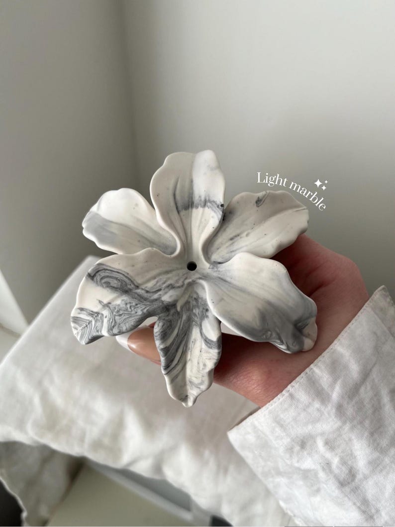 May include: A flower-shaped incense holder with a white and grey marbled pattern. The object features six curved petals and a small circular hole in the center for incense sticks. The text "Light marble" is printed in white near the top right.