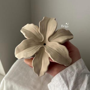 May include: A beige, flower-shaped object with six sculpted petals and a small circular opening in the center. The surface has a smooth, matte finish, and the text "Beige" with two small star icons is printed near the upper right petal.