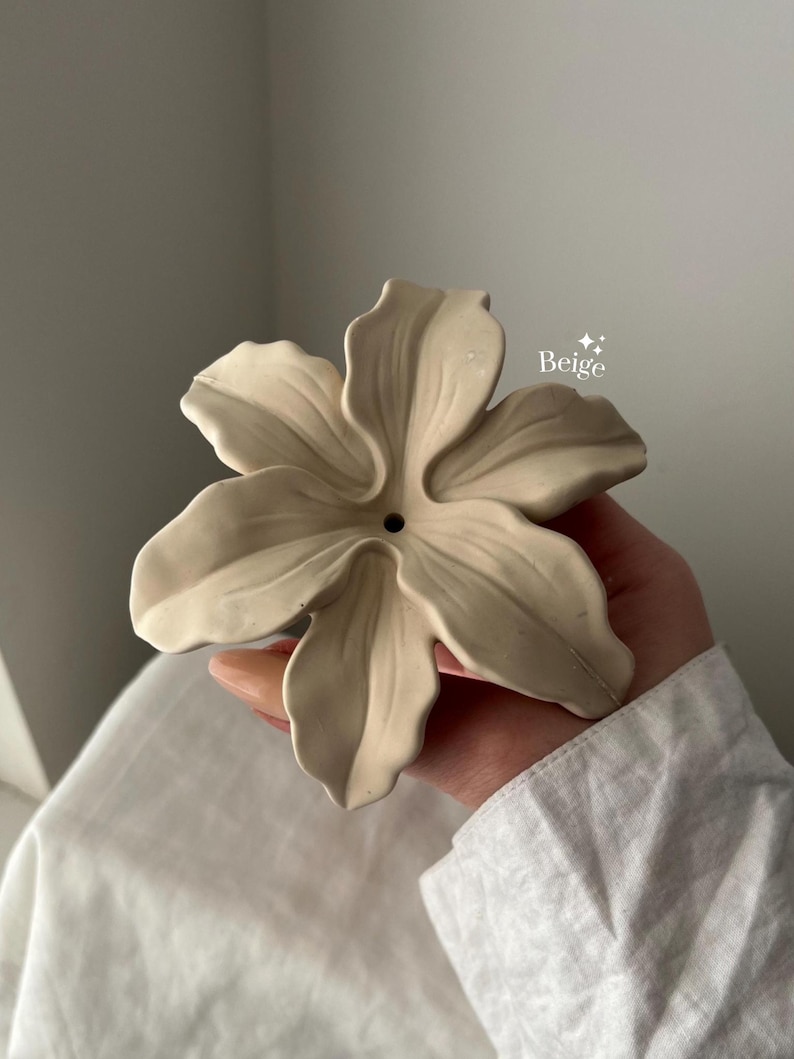 May include: A beige, flower-shaped object with six sculpted petals and a small circular opening in the center. The surface has a smooth, matte finish, and the text "Beige" with two small star icons is printed near the upper right petal.