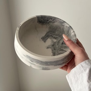 May include: A round, white and gray marbled bowl. The bowl has a smooth, polished finish with a swirling pattern of gray and white. The bowl is held by a person wearing a white shirt.