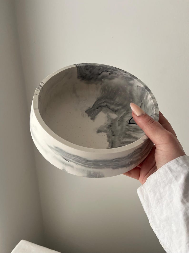 May include: A round, white and gray marbled bowl. The bowl has a smooth, polished finish with a swirling pattern of gray and white. The bowl is held by a person wearing a white shirt.