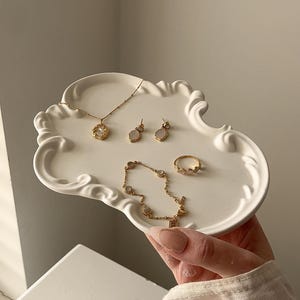 May include: A selection of gold-coloured jewellery, including a necklace, earrings, bracelet, and ring, presented on a white, ornate tray. The jewellery features delicate designs and small, light-coloured stones. The tray is held by a person.