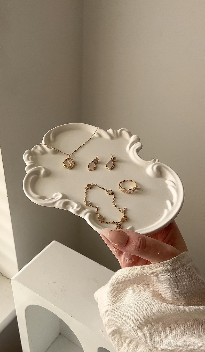 May include: A selection of gold-coloured jewellery, including a necklace, earrings, bracelet, and ring, presented on a white, ornate tray. The jewellery features delicate designs and small, light-coloured stones. The tray is held by a person.