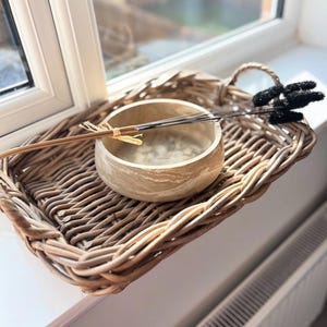 May include: A woven wicker tray with a handle holds a light-colored, textured bowl and decorative items. The bowl contains thin, dark sticks and a bundle of dried stems. A black, fluffy decorative element rests on the tray.