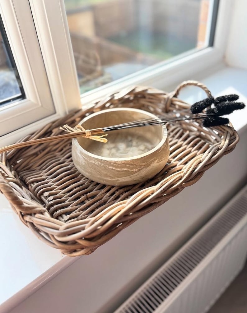 May include: A woven wicker tray with a handle holds a light-colored, textured bowl and decorative items. The bowl contains thin, dark sticks and a bundle of dried stems. A black, fluffy decorative element rests on the tray.