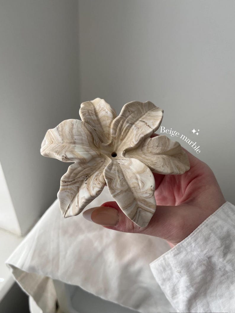 May include: A flower-shaped object with a beige marble swirl pattern. The piece features six distinct petals radiating from a central point with a small circular hole. The text "Beige marble" is printed in white on the right side of the object.