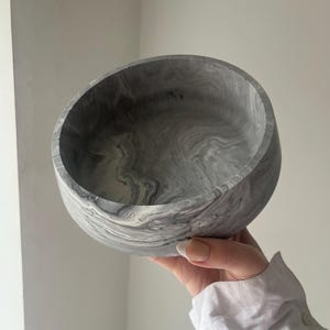 May include: A gray and white marbled bowl with a rounded shape. The bowl has a smooth, polished finish and a swirling pattern of gray and white. The bowl is held up by a person.