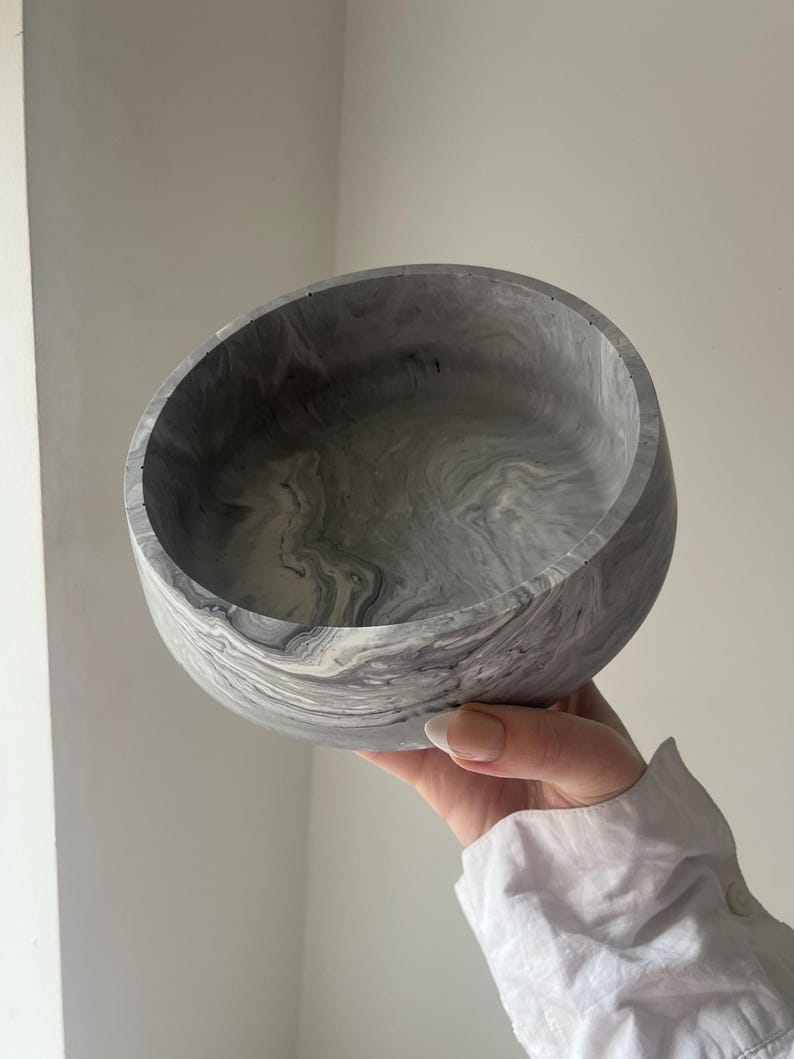May include: A gray and white marbled bowl with a rounded shape. The bowl has a smooth, polished finish and a swirling pattern of gray and white. The bowl is held up by a person.