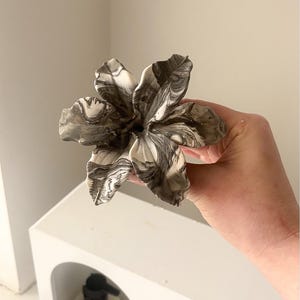 May include: A sculpted flower ornament featuring a marbled pattern of black, white, and gray swirls. The flower has six distinct, curved petals that radiate from a central point, creating a layered and organic appearance with a smooth finish.