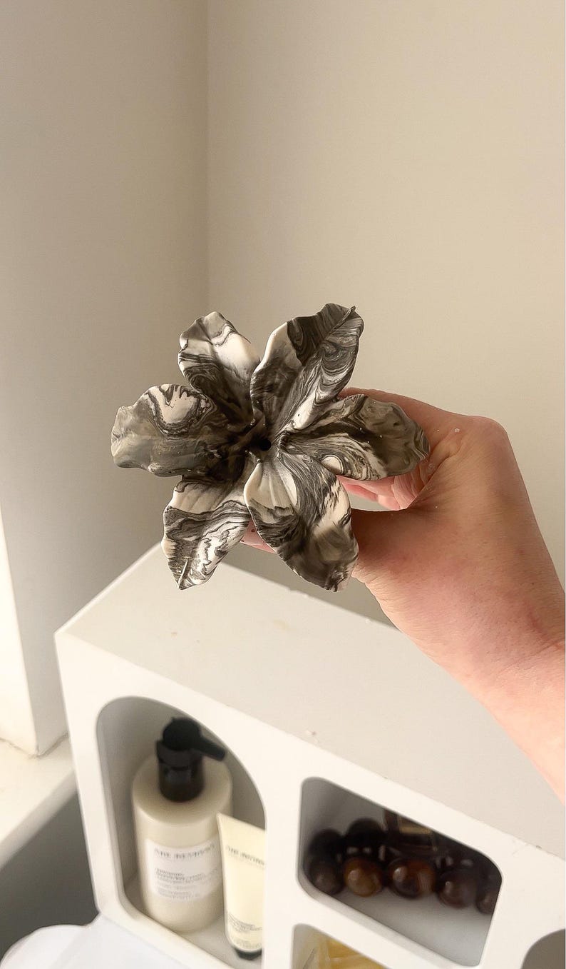 May include: A sculpted flower ornament featuring a marbled pattern of black, white, and gray swirls. The flower has six distinct, curved petals that radiate from a central point, creating a layered and organic appearance with a smooth finish.