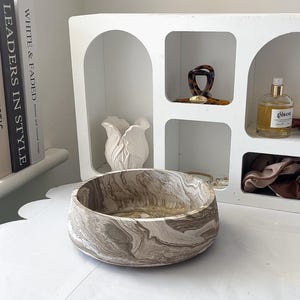 May include: A decorative, round bowl with a marbled pattern in shades of brown and cream. The bowl sits on a white surface, with a white shelf unit and books in the background. The bowl's design gives it a natural, organic feel.