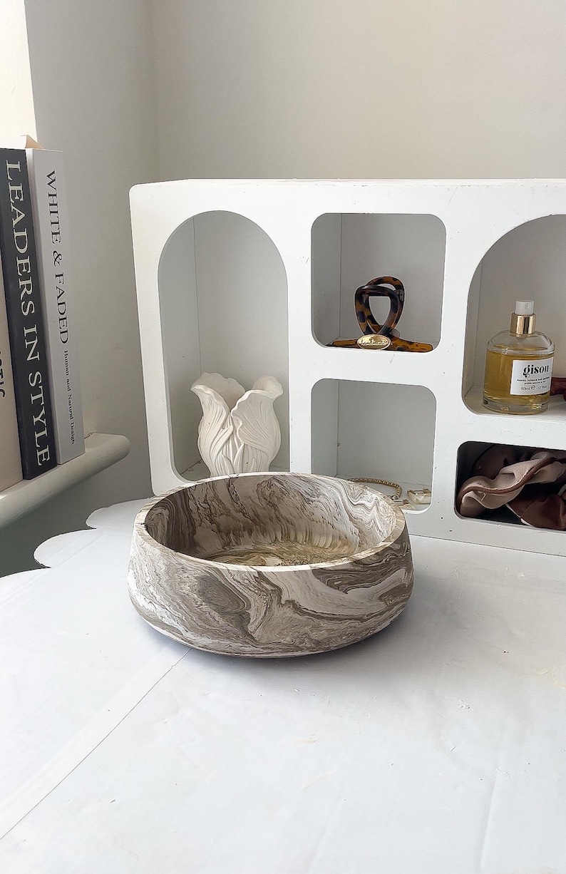 May include: A decorative, round bowl with a marbled pattern in shades of brown and cream. The bowl sits on a white surface, with a white shelf unit and books in the background. The bowl's design gives it a natural, organic feel.