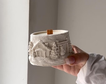 Pode incluir: Vela branca em um recipiente decorativo com uma cena de vila de inverno. O recipiente apresenta uma casa, árvores e um boneco de neve. A vela tem um pavio de madeira.