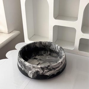 May include: A round, black and white marbled bowl. The bowl has a black base and a swirling pattern of white and gray. The bowl is sitting on a white surface, with a white shelf unit in the background.