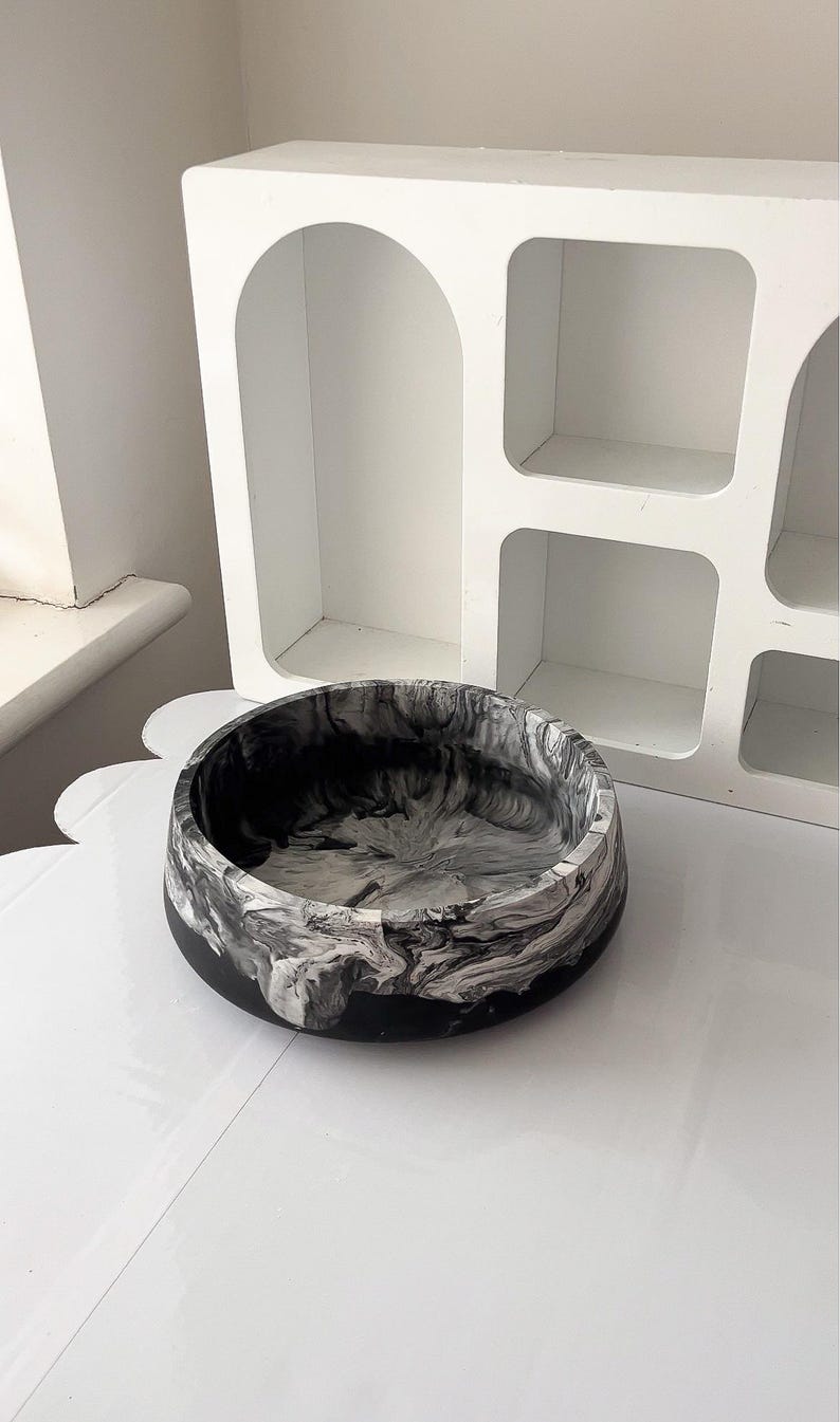 May include: A round, black and white marbled bowl. The bowl has a black base and a swirling pattern of white and gray. The bowl is sitting on a white surface, with a white shelf unit in the background.