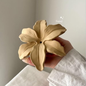 May include: A sand-colored, flower-shaped object with six petals and a small circular hole in the center. The surface has a matte finish with subtle texture. The word Sand is printed in white text above the flower, accompanied by two small star icons.