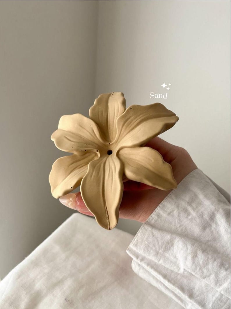 May include: A sand-colored, flower-shaped object with six petals and a small circular hole in the center. The surface has a matte finish with subtle texture. The word Sand is printed in white text above the flower, accompanied by two small star icons.